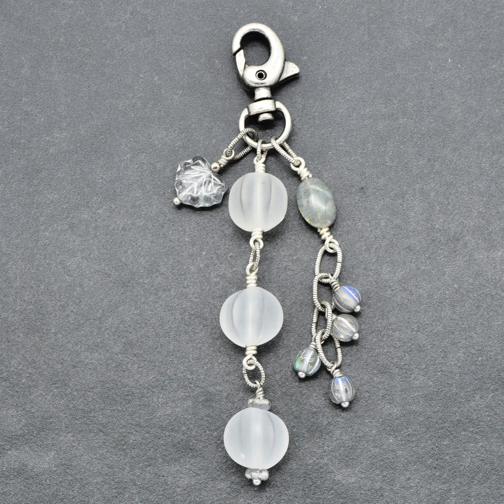 Snowfall Serenade handmade bag charm with three frosted white fluted Lucite beads, a labradorite oval, gray Czech glass beads, a clear glass maple leaf, and an antique-silver-finish swivel clasp on a gray background.