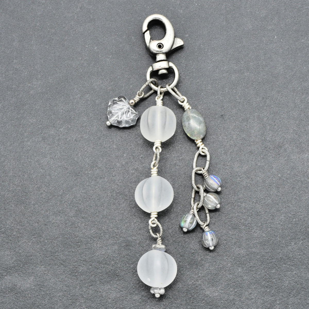 Snowfall Serenade handmade bag charm with three frosted white fluted Lucite beads, a labradorite oval, gray Czech glass beads, a clear glass maple leaf, and an antique-silver-finish swivel clasp on a gray background.