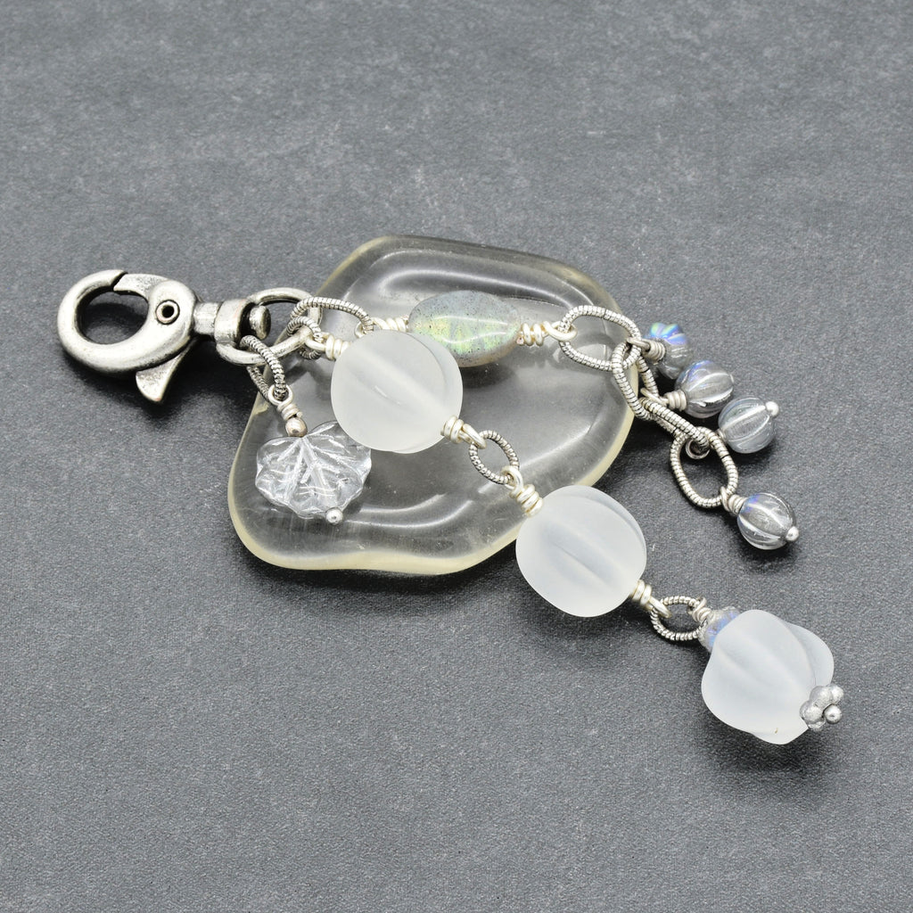Snowfall Serenade handmade bag charm with three frosted white fluted Lucite beads, a labradorite oval, gray Czech glass beads, a clear glass maple leaf, and an antique-silver-finish swivel clasp on a gray background.