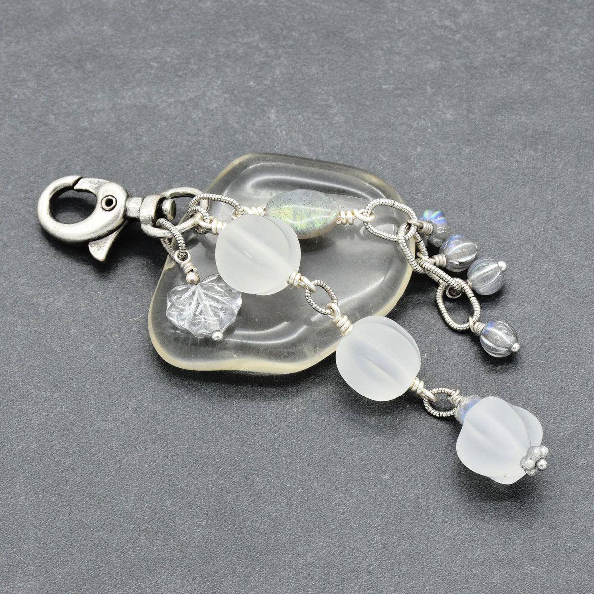 Snowfall Serenade handmade bag charm with three frosted white fluted Lucite beads, a labradorite oval, gray Czech glass beads, a clear glass maple leaf, and an antique-silver-finish swivel clasp on a gray background.