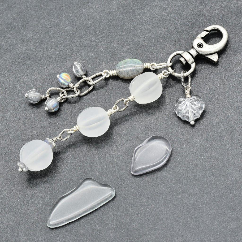Snowfall Serenade handmade bag charm with three frosted white fluted Lucite beads, a labradorite oval, gray Czech glass beads, a clear glass maple leaf, and an antique-silver-finish swivel clasp on a gray background.