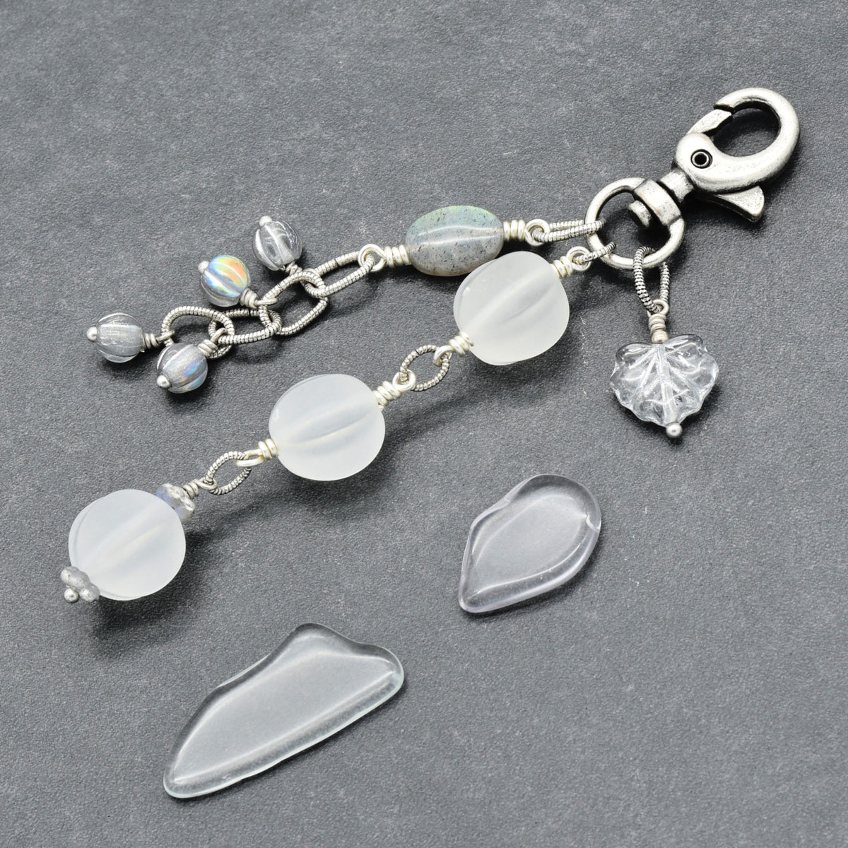 Snowfall Serenade handmade bag charm with three frosted white fluted Lucite beads, a labradorite oval, gray Czech glass beads, a clear glass maple leaf, and an antique-silver-finish swivel clasp on a gray background.