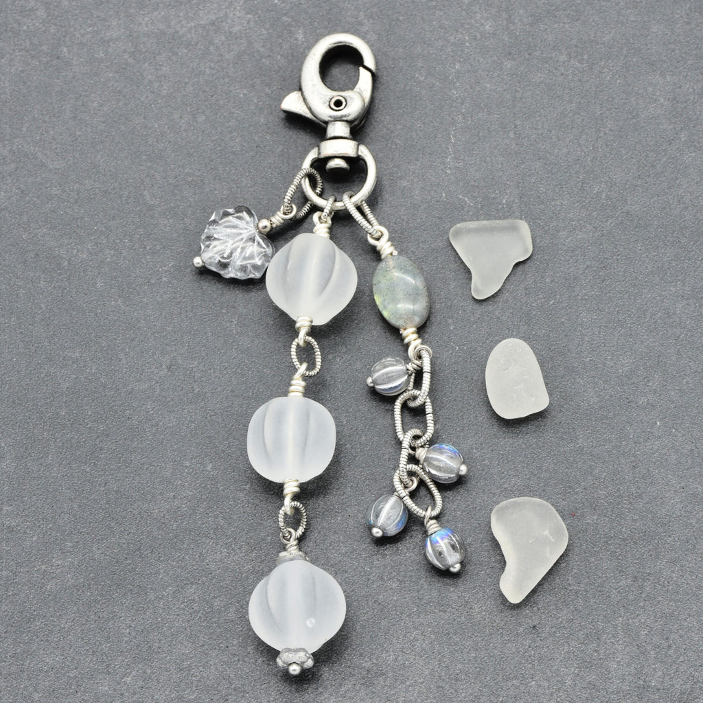 Snowfall Serenade handmade bag charm with three frosted white fluted Lucite beads, a labradorite oval, gray Czech glass beads, a clear glass maple leaf, and an antique-silver-finish swivel clasp on a gray background.