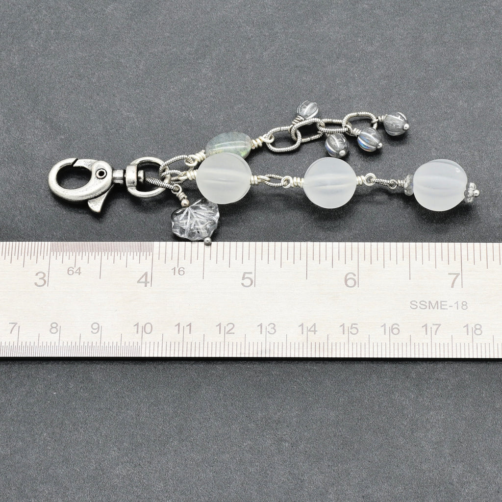 Snowfall Serenade handmade bag charm with three frosted white fluted Lucite beads, a labradorite oval, gray Czech glass beads, a clear glass maple leaf, and an antique-silver-finish swivel clasp next to a ruler for scale.