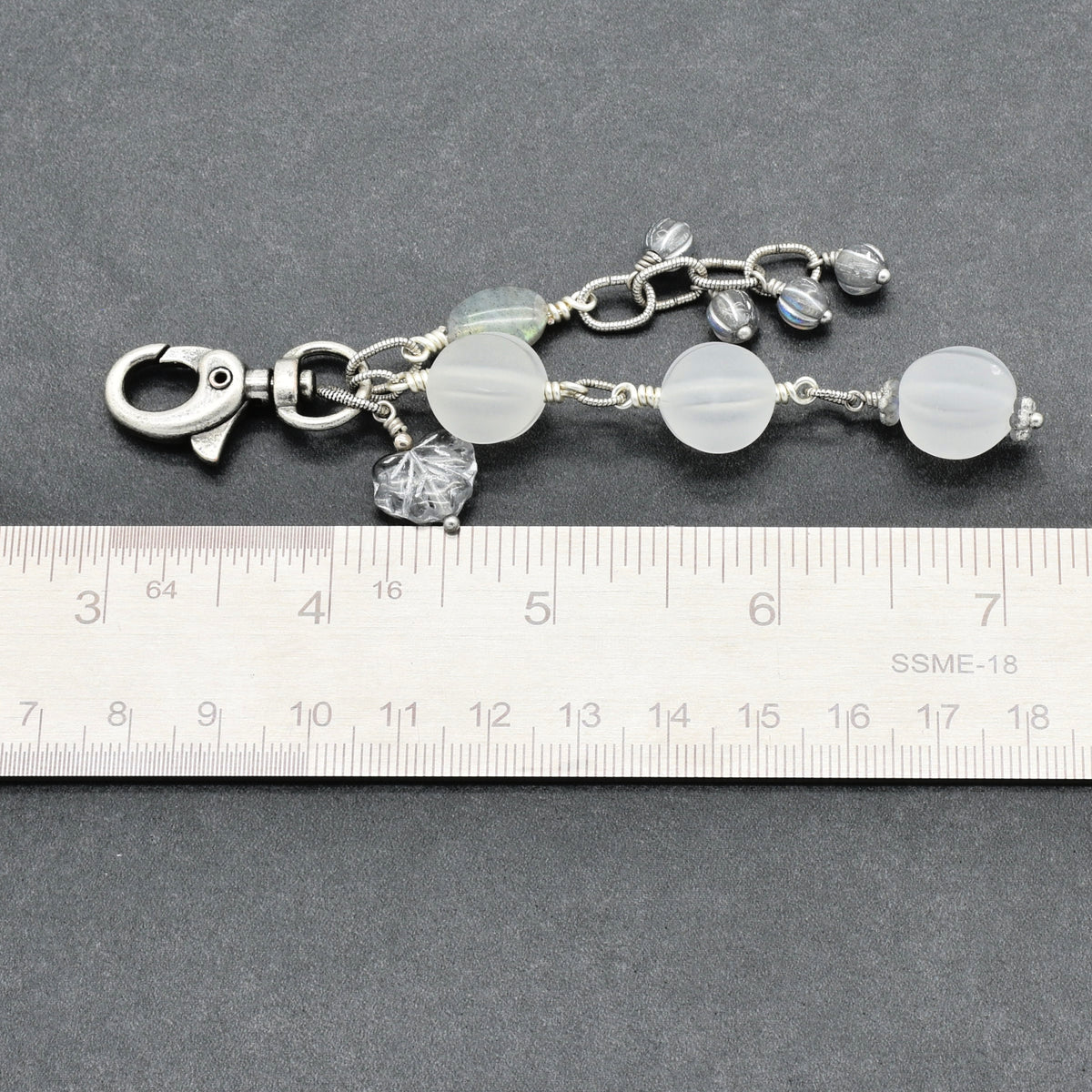 Snowfall Serenade handmade bag charm with three frosted white fluted Lucite beads, a labradorite oval, gray Czech glass beads, a clear glass maple leaf, and an antique-silver-finish swivel clasp next to a ruler for scale.