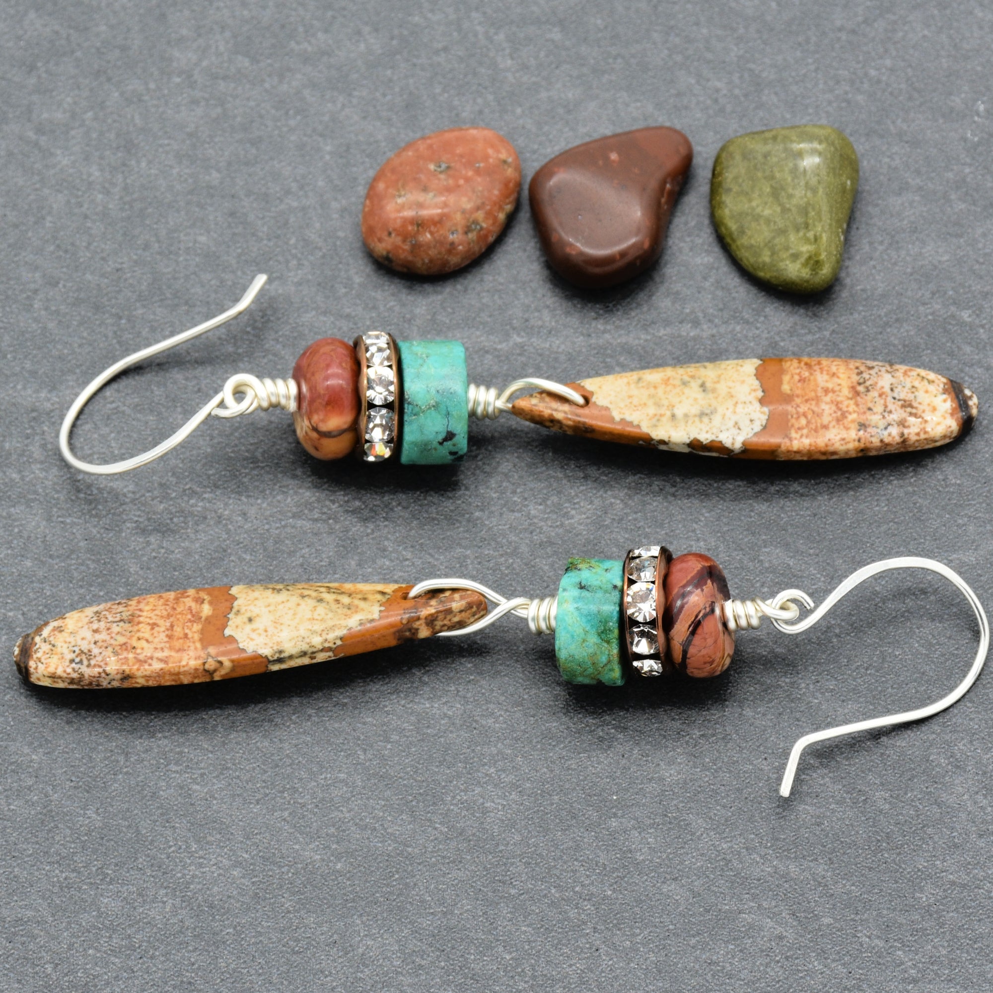 The jewelry in the image is a pair of earrings. They feature a combination of earthy-toned beads, including a long, oval-shaped bead, turquoise-colored beads, and accents with small clear rhinestones. The earrings have hook-style ear wires for wearing.