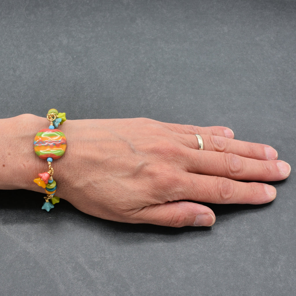 Artist's hand wearing happy colorful bracelet with African Kazuri focal, tagua nuts, and Czech glass flowers in tangerine, lime, marigold and turquoise on a light surface - handmade with love by Ruby Acorn