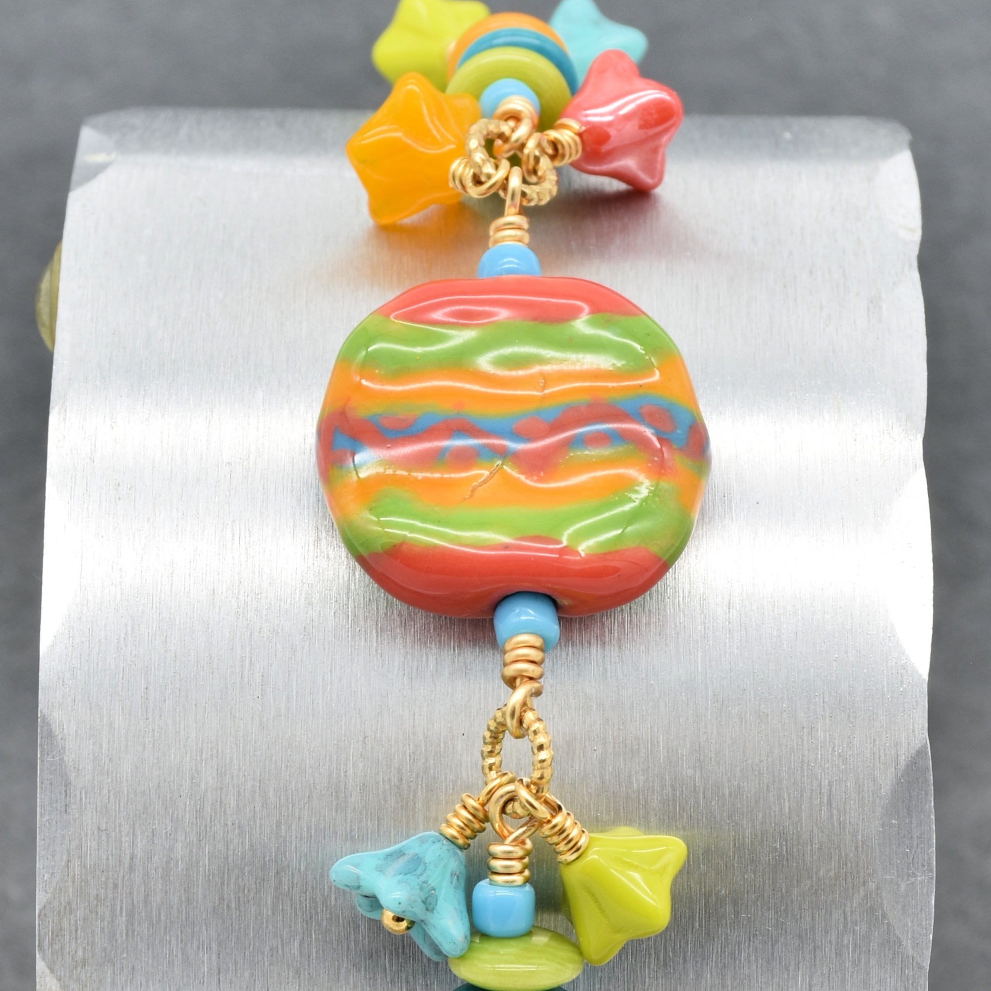 Closeup of happy colorful bracelet with African Kazuri focal, tagua nuts, and Czech glass flowers in tangerine, lime, marigold and turquoise on a silver surface - handmade with love by Ruby Acorn