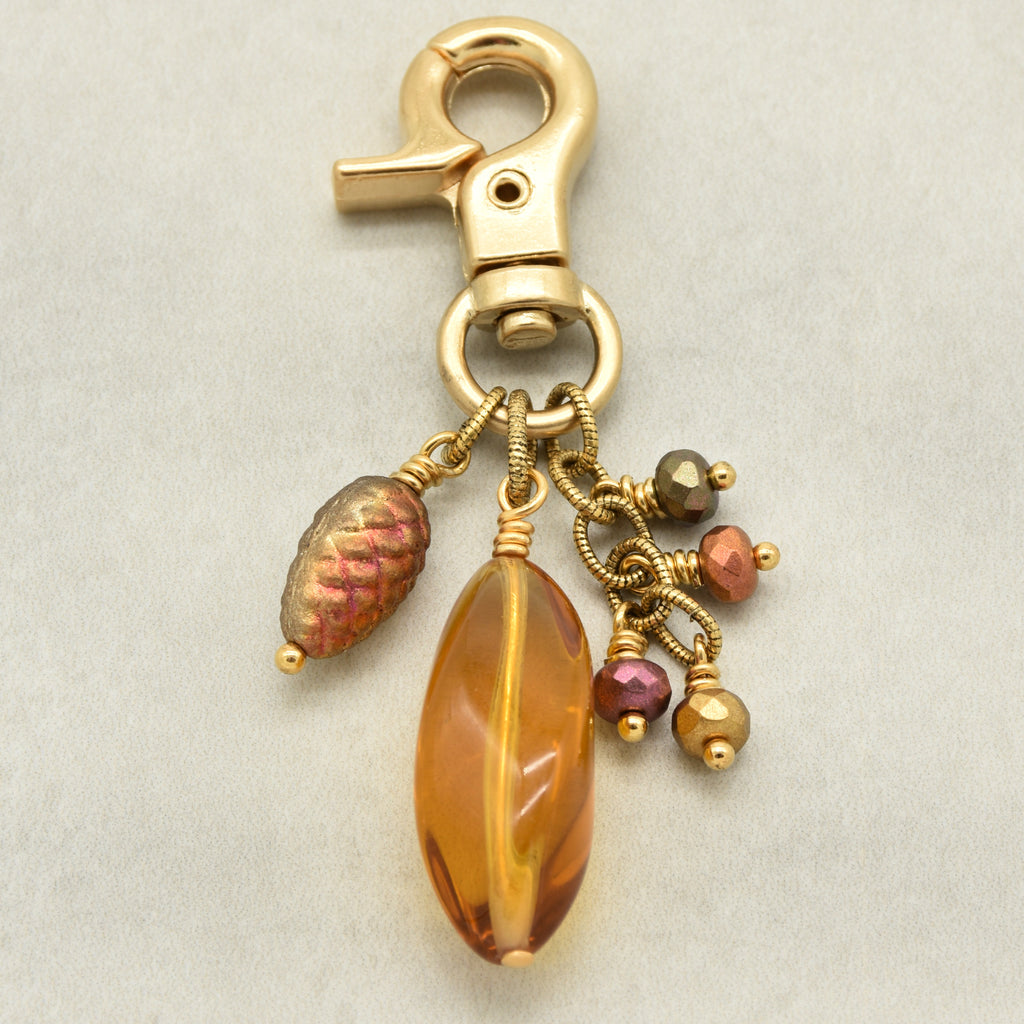 A gold-toned lobster clasp keychain charm featuring a large amber Lucite bead, a textured metallic pinecone bead, and small faceted beads in pink, purple, and brown - made with love in Colorado by Ruby Acorn.
