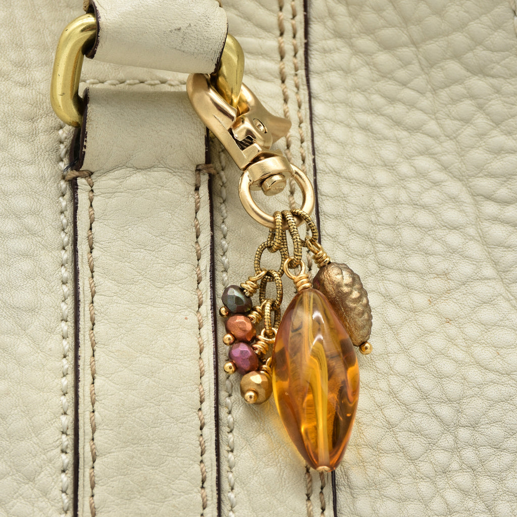 Close-up of a cream-colored leather bag featuring a decorative gold-tone clasp and charm with a large amber-colored faceted bead and small multicolor glass beads - made with love in Colorado by Ruby Acorn.