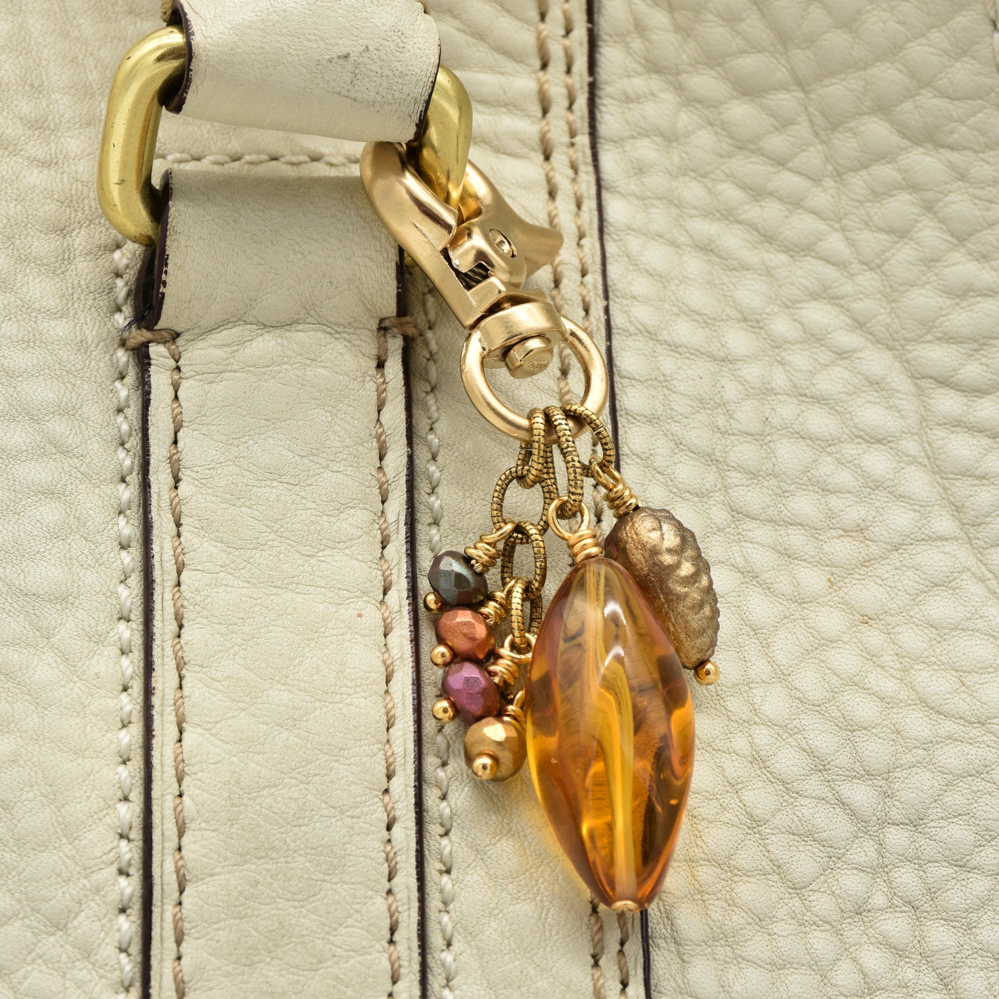 Close-up of a cream-colored leather bag featuring a decorative gold-tone clasp and charm with a large amber-colored faceted bead and small multicolor glass beads - made with love in Colorado by Ruby Acorn.