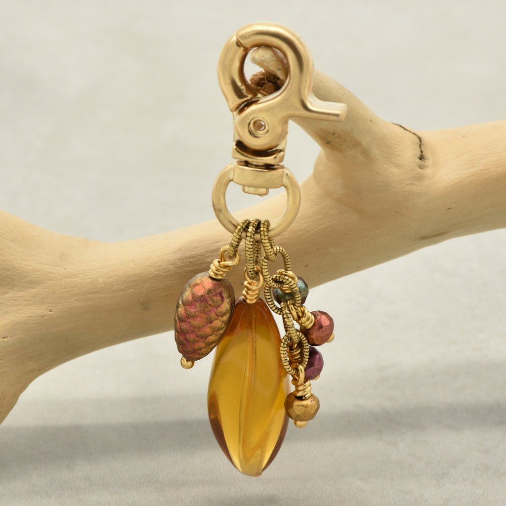 A gold-toned lobster clasp keychain or bag charm hanging from a light wood branch. The charm features a large amber-yellow Lucite teardrop bead, a textured iridescent pinecone bead, and small colorful accent beads - made with love in Colorado by Ruby Acorn.