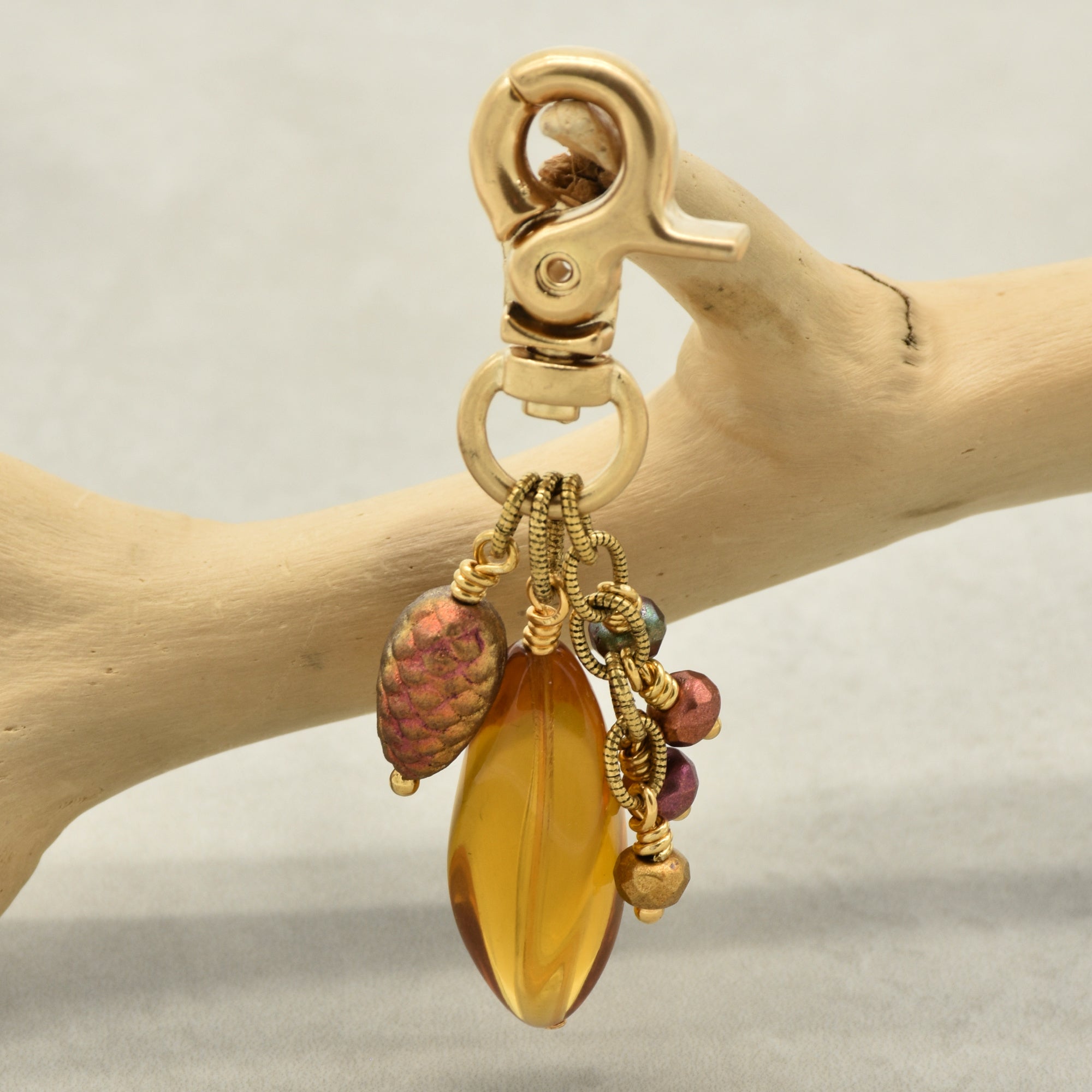 A gold-toned lobster clasp keychain or bag charm hanging from a light wood branch. The charm features a large amber-yellow Lucite teardrop bead, a textured iridescent pinecone bead, and small colorful accent beads - made with love in Colorado by Ruby Acorn.