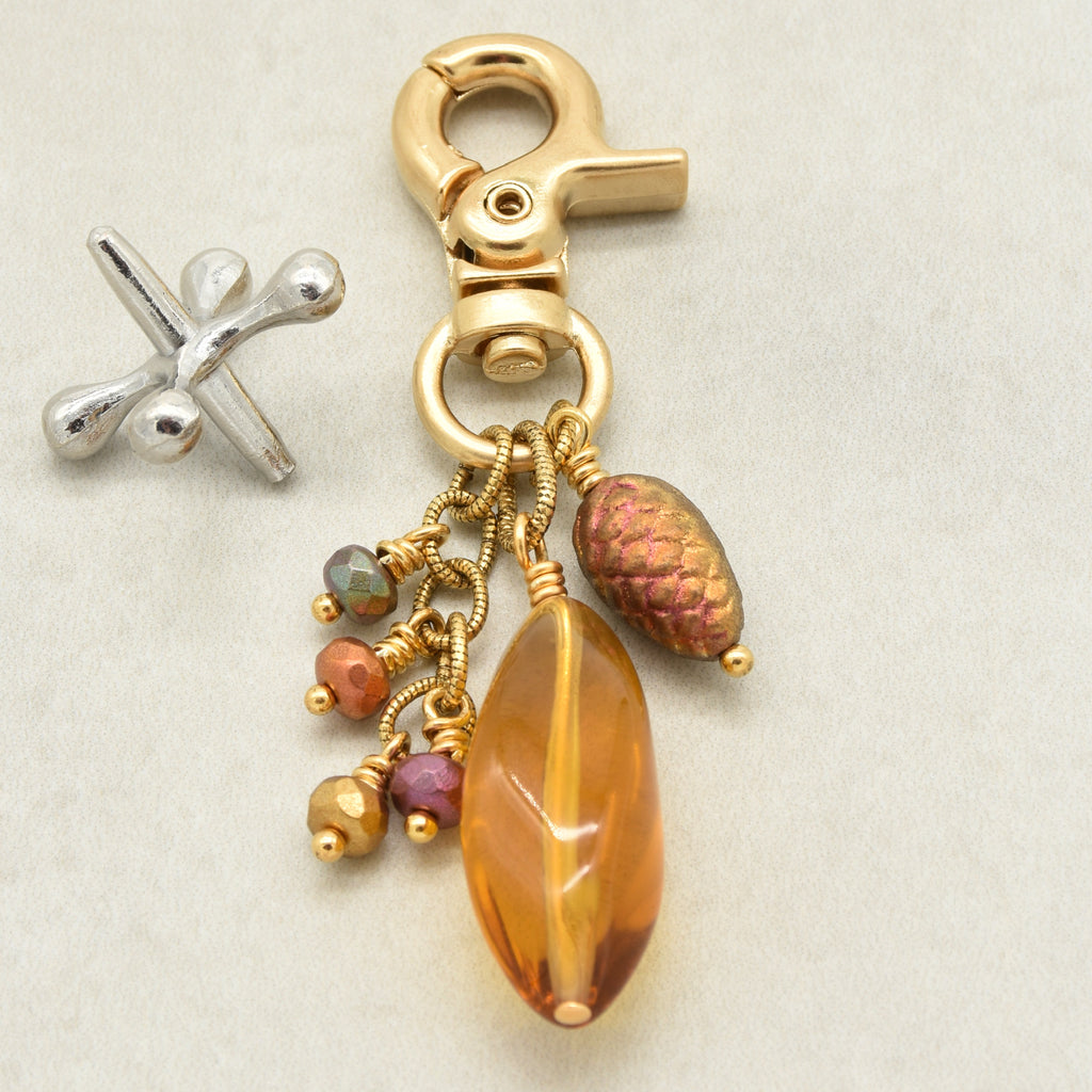A gold-toned lobster clasp keychain charm with a large faceted amber Lucite bead, a textured pinecone bead, and small multicolored beads, shown on a light beige background - made with love in Colorado by Ruby Acorn.