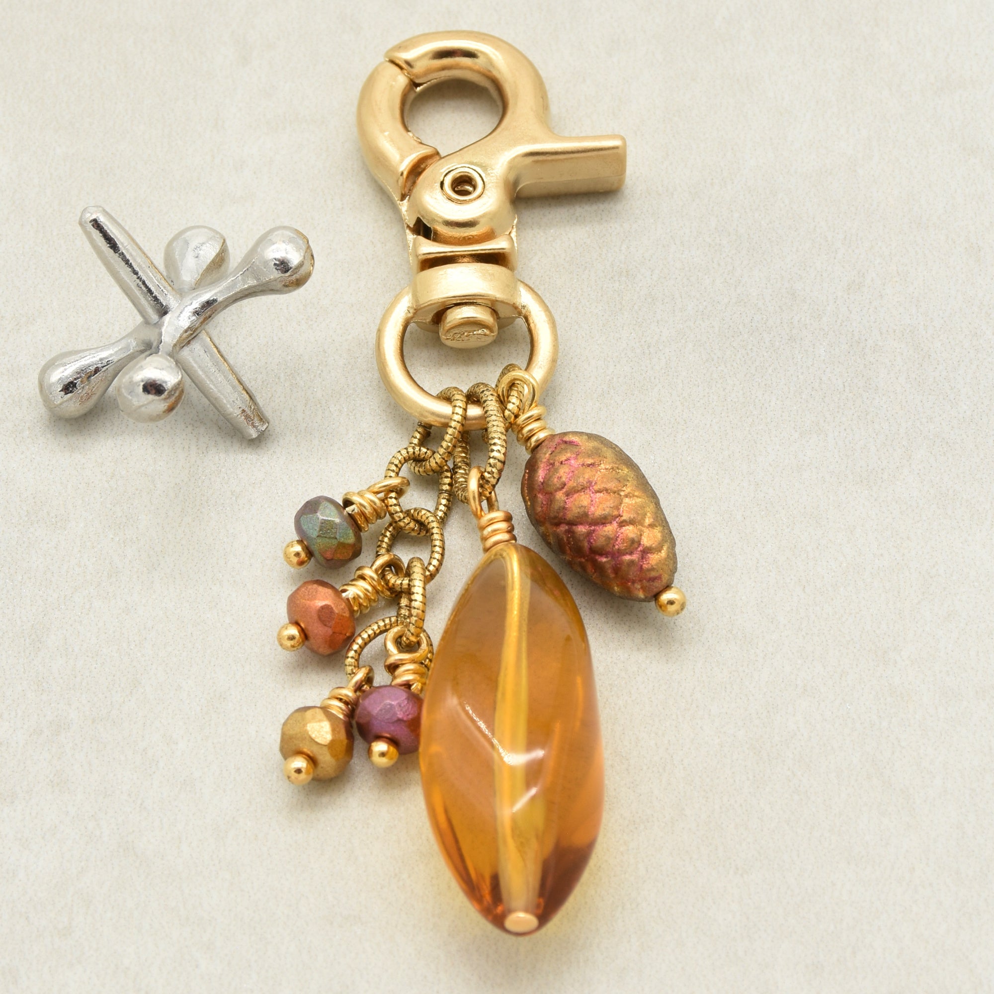 A gold-toned lobster clasp keychain charm with a large faceted amber Lucite bead, a textured pinecone bead, and small multicolored beads, shown on a light beige background - made with love in Colorado by Ruby Acorn.