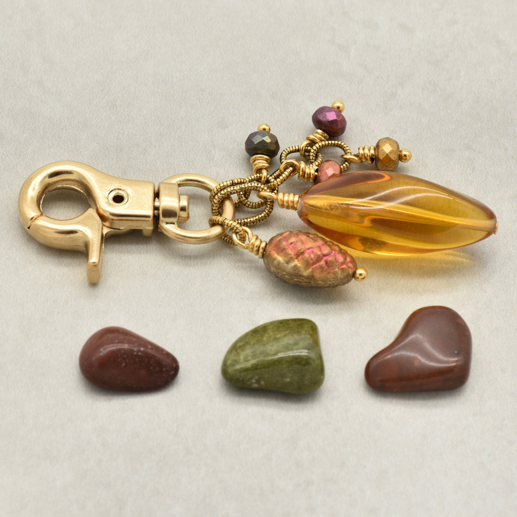 A gold-toned keychain or bag charm with a lobster clasp, amber Lucite focal, pinecone charm, and glass accents, displayed on a light gray background with three small polished stones placed below - made with love in Colorado by Ruby Acorn.