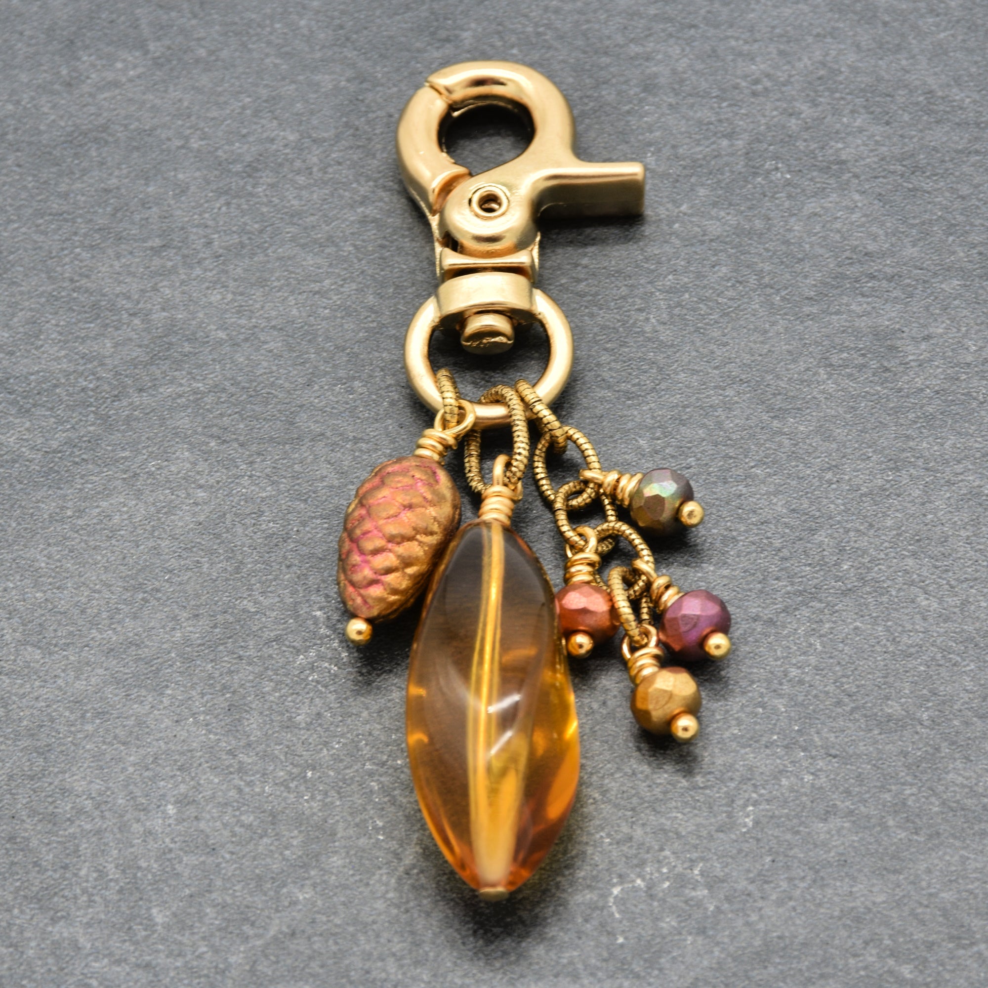 A gold-toned metal keychain or bag charm with a lobster clasp, featuring a large amber Lucite bead, a textured pinkish-brown pinecone bead, and smaller multicolored beads on gold wire, shown on a dark gray textured background - made with love in Colorado by Ruby Acorn.