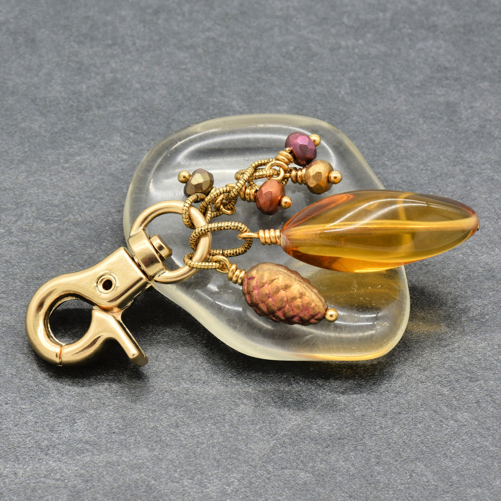 A handmade keychain or bag charm displayed on a large clear piece of glass, a polished amber-yellow Lucite teardrop bead is the focal, a textured brown pinecone bead, and small colorful accent beads complete the design, all attached to a gold-toned lobster clasp, set against a dark gray textured background - made with love in Colorado by Ruby Acorn.
