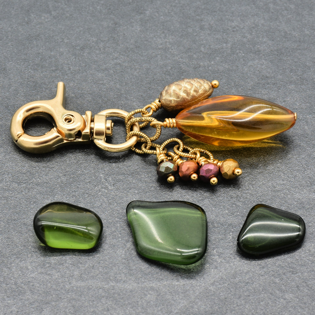A gold-tone keychain or bag charm with a lobster clasp, a gold acorn bead, a translucent amber teardrop Lucite bead, and three dangling polished glass beads, displayed on a gray textured background - made with love by Ruby Acorn in Colorado.