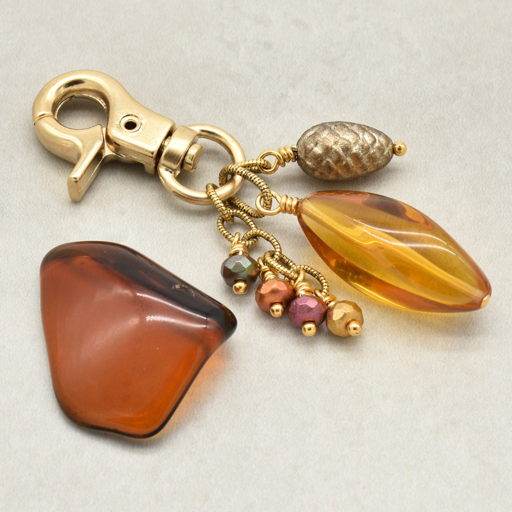 A gold-toned keychain or bag charm with a lobster clasp, featuring a large faceted amber-yellow Lucite teardrop bead, a textured metallic gold pinecone bead, and small dangling beads in amber, pink, and green - made with love in Colorado by Ruby Acorn.