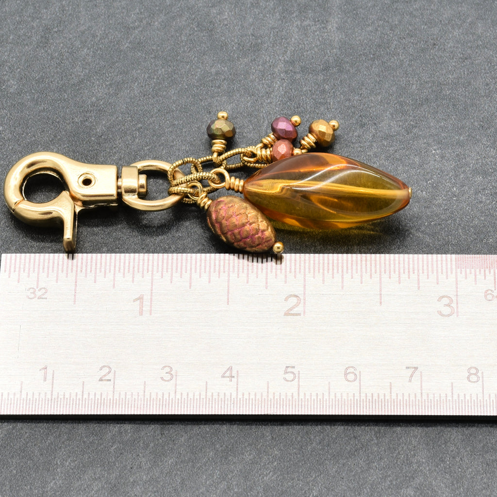 A vintage-style gold-toned keychain or bag charm with a lobster clasp, a large amber Lucite teardrop bead, a textured brown pinecone charm, and small pink and gold beads, shown next to a metal ruler on a dark gray background - made with love in Colorado by Ruby Acorn.