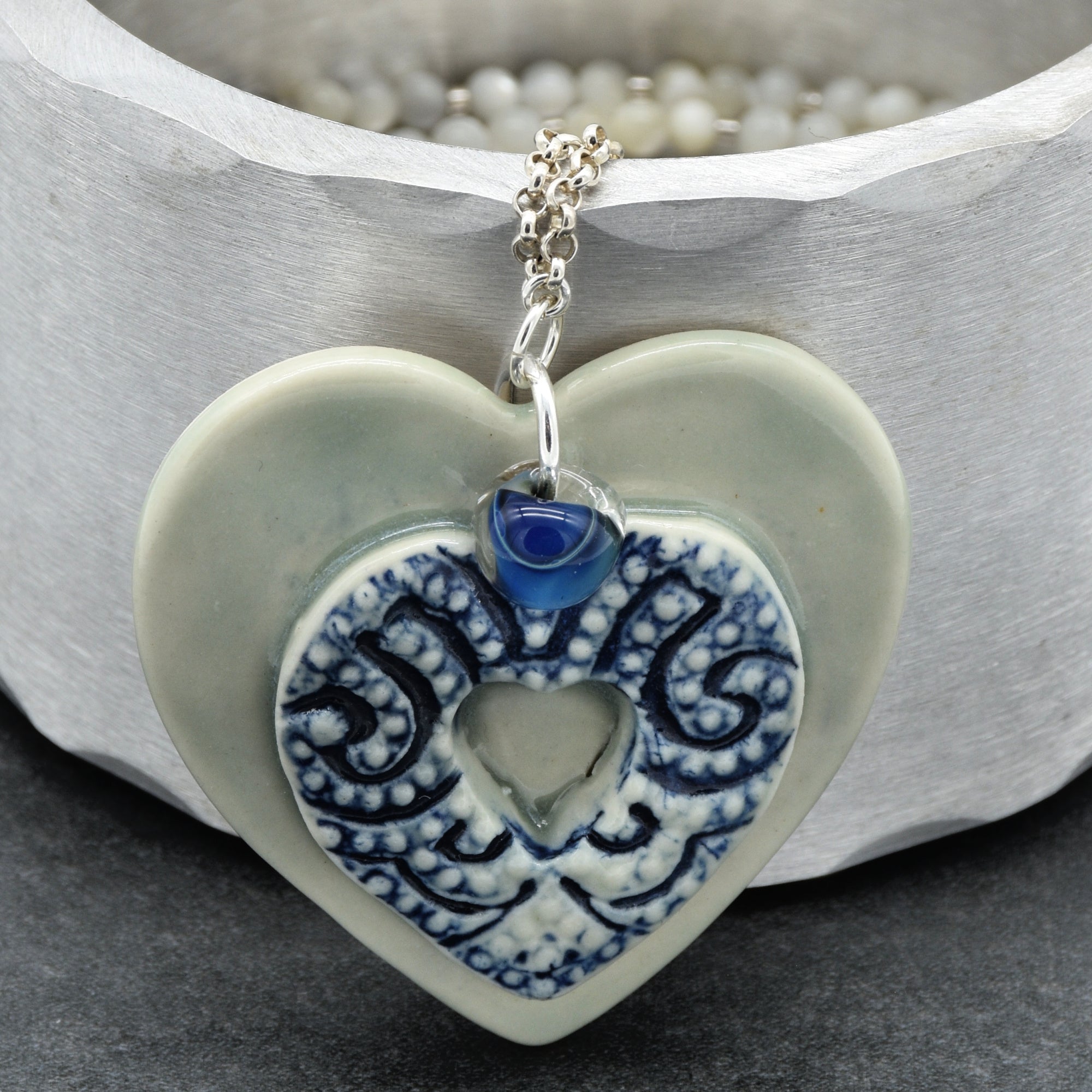 Heart-shaped ceramic pendant with blue pattern and blue crystal on a silver chain, against a neutral background.