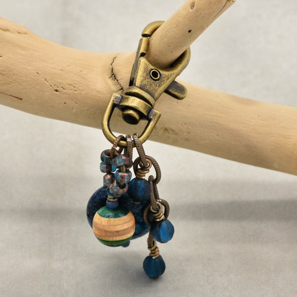 An antique bronze lobster clasp jewelry charm with blue beads, and a striped wooden bead, hanging from a light wood branch against a neutral gray background - made with love in Colorado by Ruby Acorn.