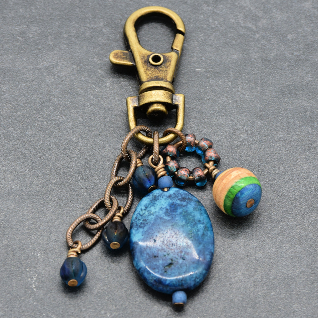 A vintage-style keychain or bag charm featuring an antique bronze-toned lobster clasp, a large polished blue stone bead, smaller blue beads, wrapped antique brass wire charms, and a striped wooden bead on a dark gray textured background - made with love in Colorado by Ruby Acorn.