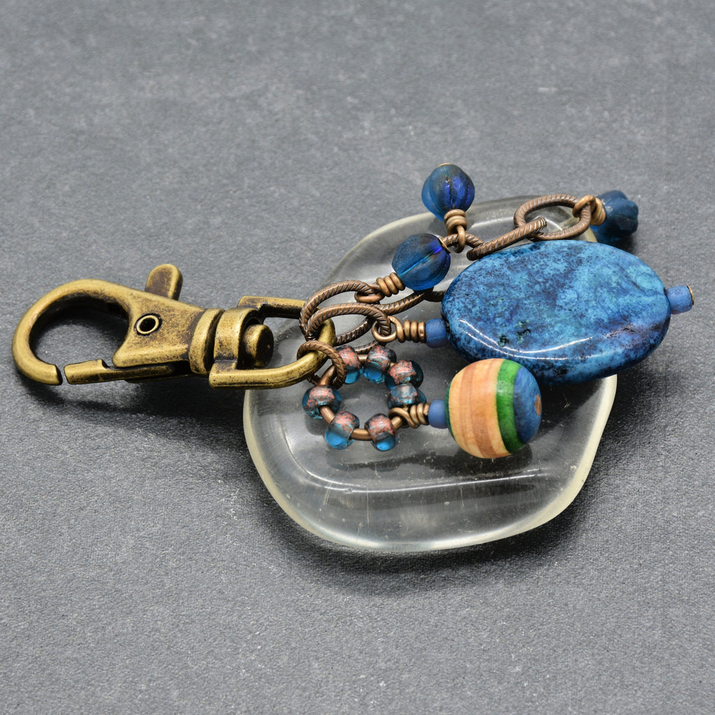 A handmade keychain featuring a bronze-finished lobster clasp, a large blue stone bead, a striped wooden bead, and small blue beads on a clear glass stand against a dark gray background - made with love in Colorado by Ruby Acorn.