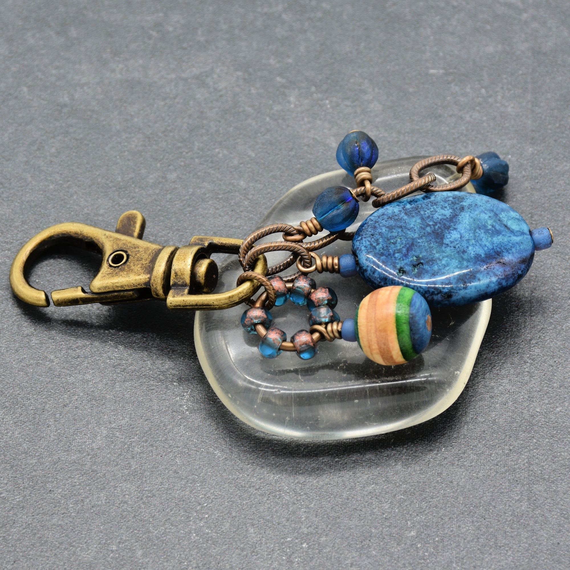 A handmade keychain featuring a bronze-finished lobster clasp, a large blue stone bead, a striped wooden bead, and small blue beads on a clear glass stand against a dark gray background - made with love in Colorado by Ruby Acorn.