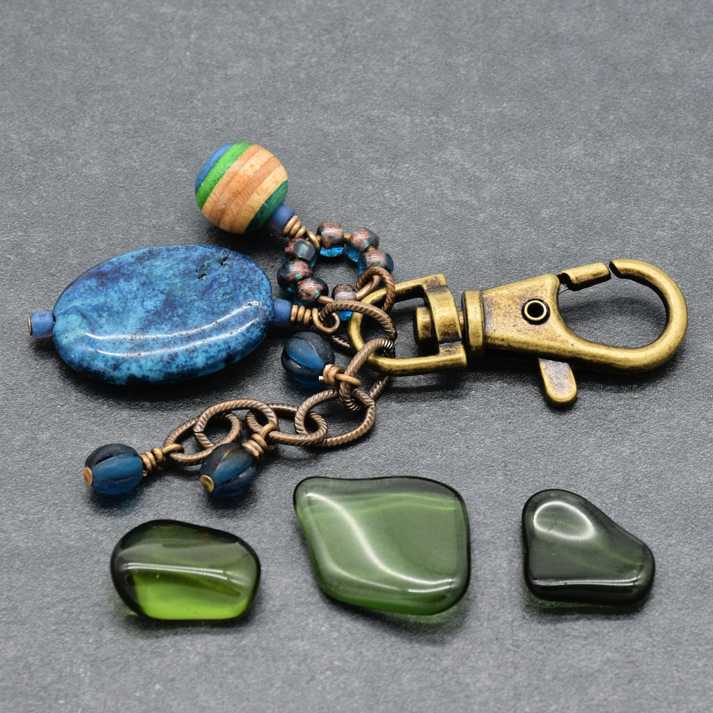 A handmade keychain or bag charm with a blue stone, bronze-toned metal chain, a striped wooden bead, and three green tumbled stones at props on a gray textured background - made with love in Colorado by Ruby Acorn.