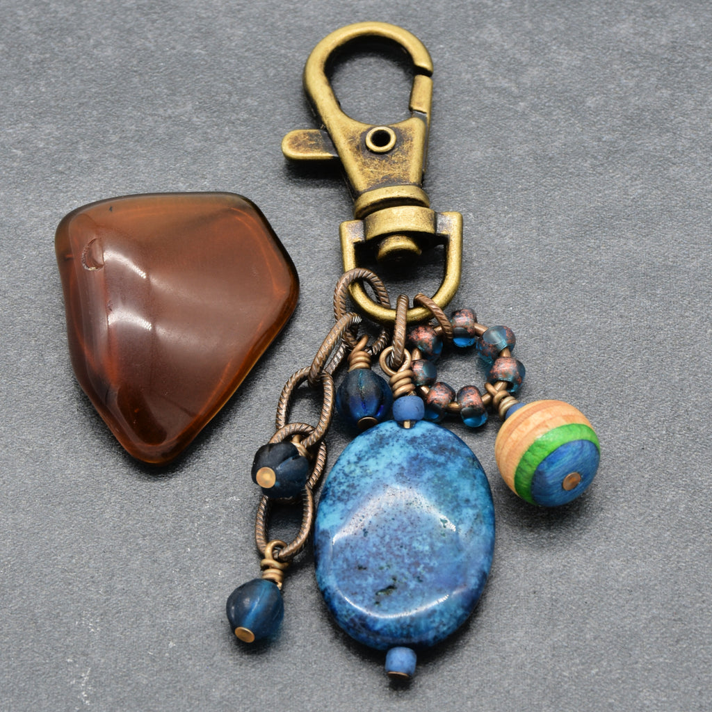 A vintage-style jewelry keychain or bag charm with an antique brass clasp, a large blue agate bead, blue beads, a striped wooden bead, and a polished brown stone as a prop, displayed on a gray textured background - made with love in Colorado by Ruby Acorn.