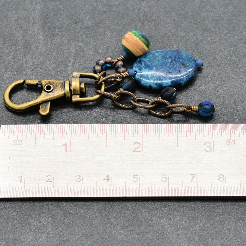 A handmade keychain or bag charm featuring a brass-colored lobster clasp, a bronze chain, a blue stone bead, a striped wooden bead, and small dark beads, shown next to a metal ruler for scale on a gray background - made with love in Colorado by Ruby Acorn.