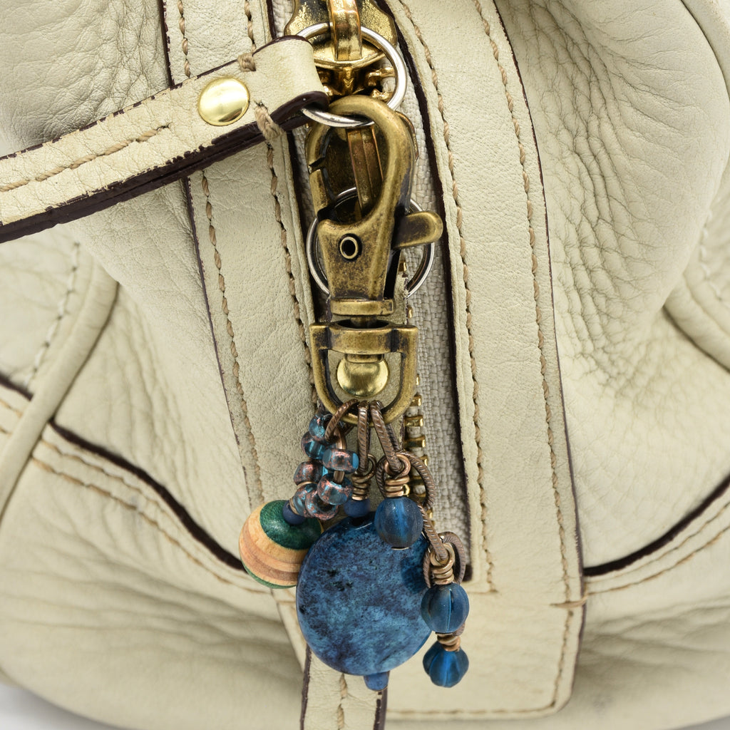 Close-up of a cream-colored textured leather handbag featuring a brass-tone lobster clasp and a decorative hanging charm with blue beads and wooden accents - made with love in Colorado by Ruby Acorn.