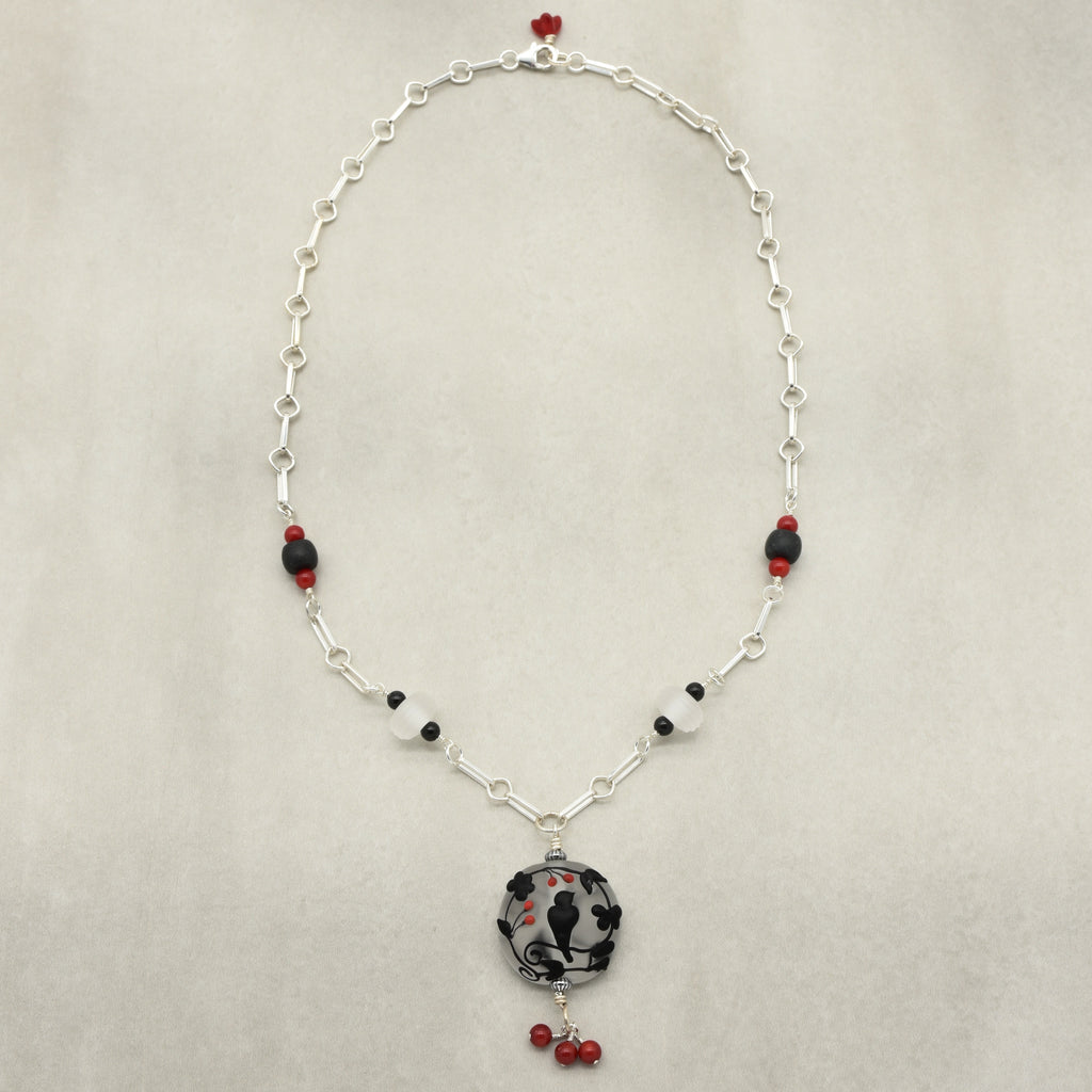 A silver link chain necklace featuring a central round black and white patterned glass bead pendant, accented with small red and black beads on a light beige background - made with love in Colorado by Ruby Acorn.