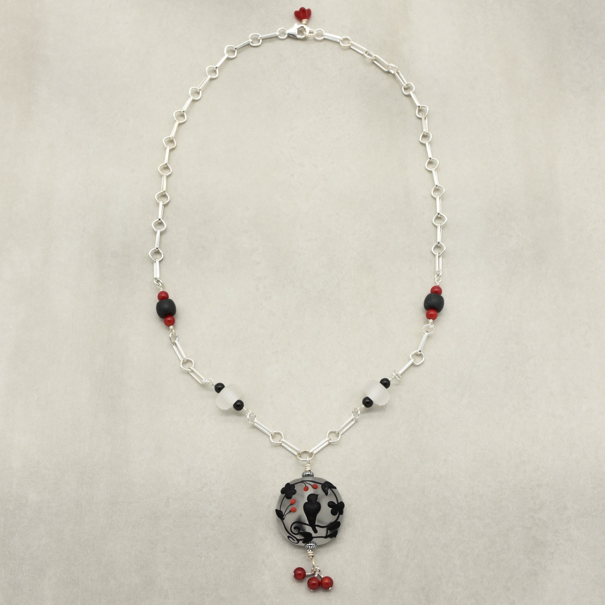 A silver link chain necklace featuring a central round black and white patterned glass bead pendant, accented with small red and black beads on a light beige background - made with love in Colorado by Ruby Acorn.