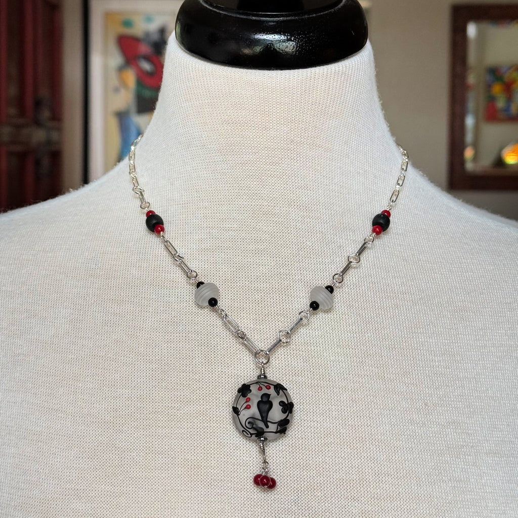 A silver-toned beaded necklace displayed on a white fabric mannequin bust. The necklace features a central round pendant with a black and white floral pattern and small red accents, accented by white beads, black beads, and silver chain links - made with love in Colorado by Ruby Acorn.