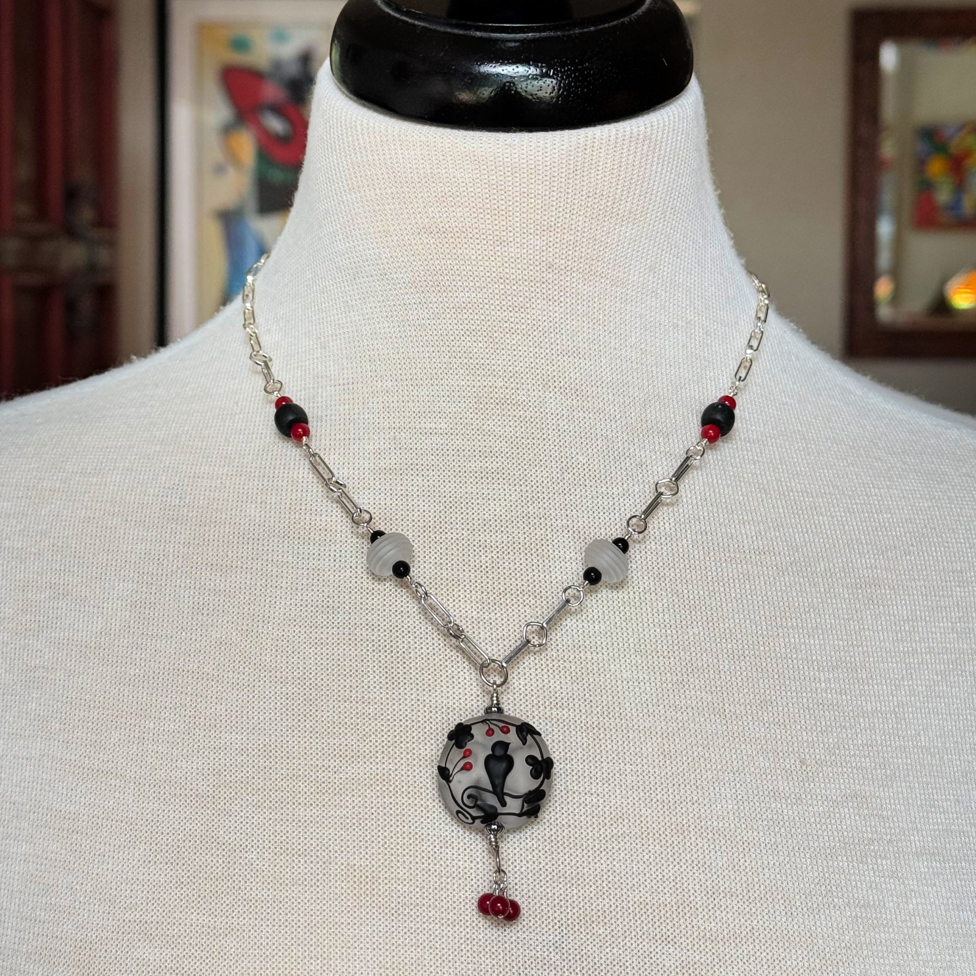 A silver-toned beaded necklace displayed on a white fabric mannequin bust. The necklace features a central round pendant with a black and white floral pattern and small red accents, accented by white beads, black beads, and silver chain links - made with love in Colorado by Ruby Acorn.