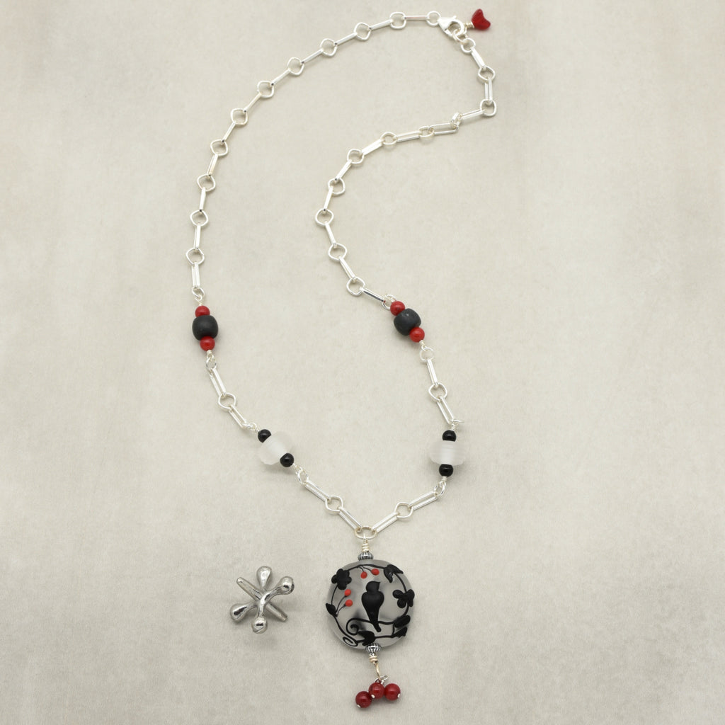A silver-toned wire wrapped necklace with a circular pendant featuring a black raven, accented with red beads, displayed on a neutral beige background - made with love in Colorado by Ruby Acorn.