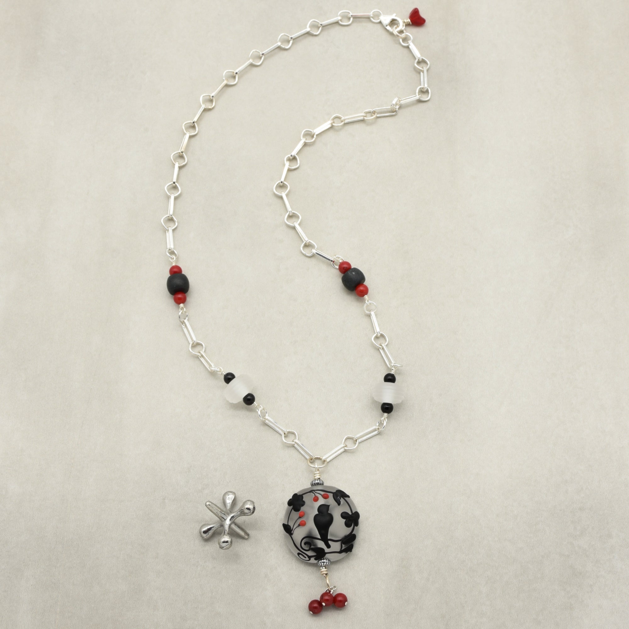 A silver-toned wire wrapped necklace with a circular pendant featuring a black raven, accented with red beads, displayed on a neutral beige background - made with love in Colorado by Ruby Acorn.