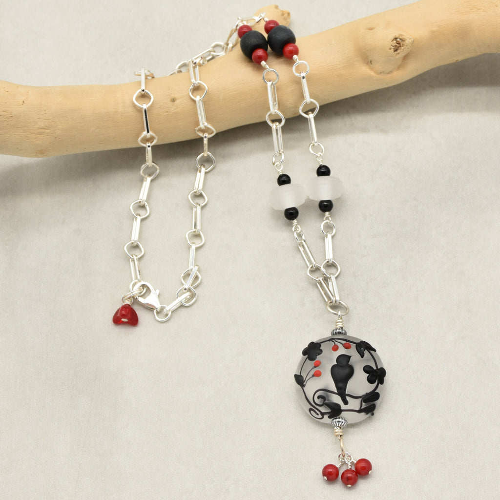 A silver link chain necklace with black and red beads and a circular black raven silhouette pendant, displayed on a light wood branch against a neutral light gray background - made with love in Colorado by Ruby Acorn.