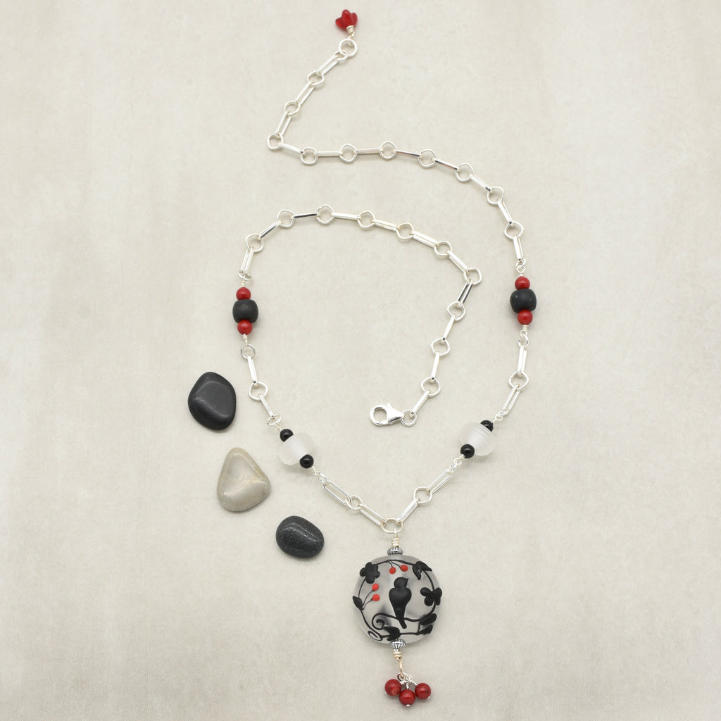A silver chain necklace with alternating white and red beads, featuring a circular pendant with a black raven at the center, displayed on a light beige background with decorative stones - made with love in Colorado by Ruby Acorn.