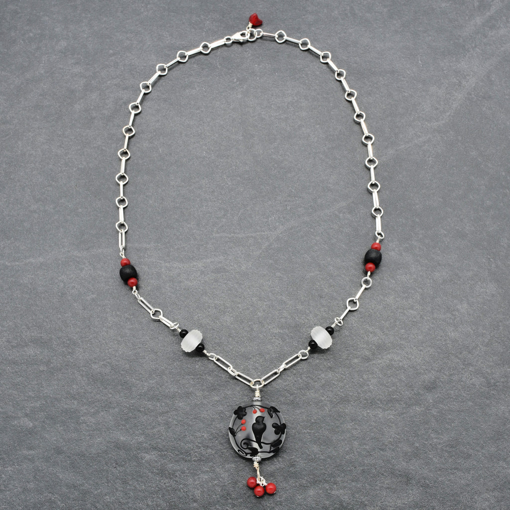 A silver-toned beaded necklace with a circular black and red pendant, displayed on a textured gray background - made with love in Colorado by Ruby Acorn.