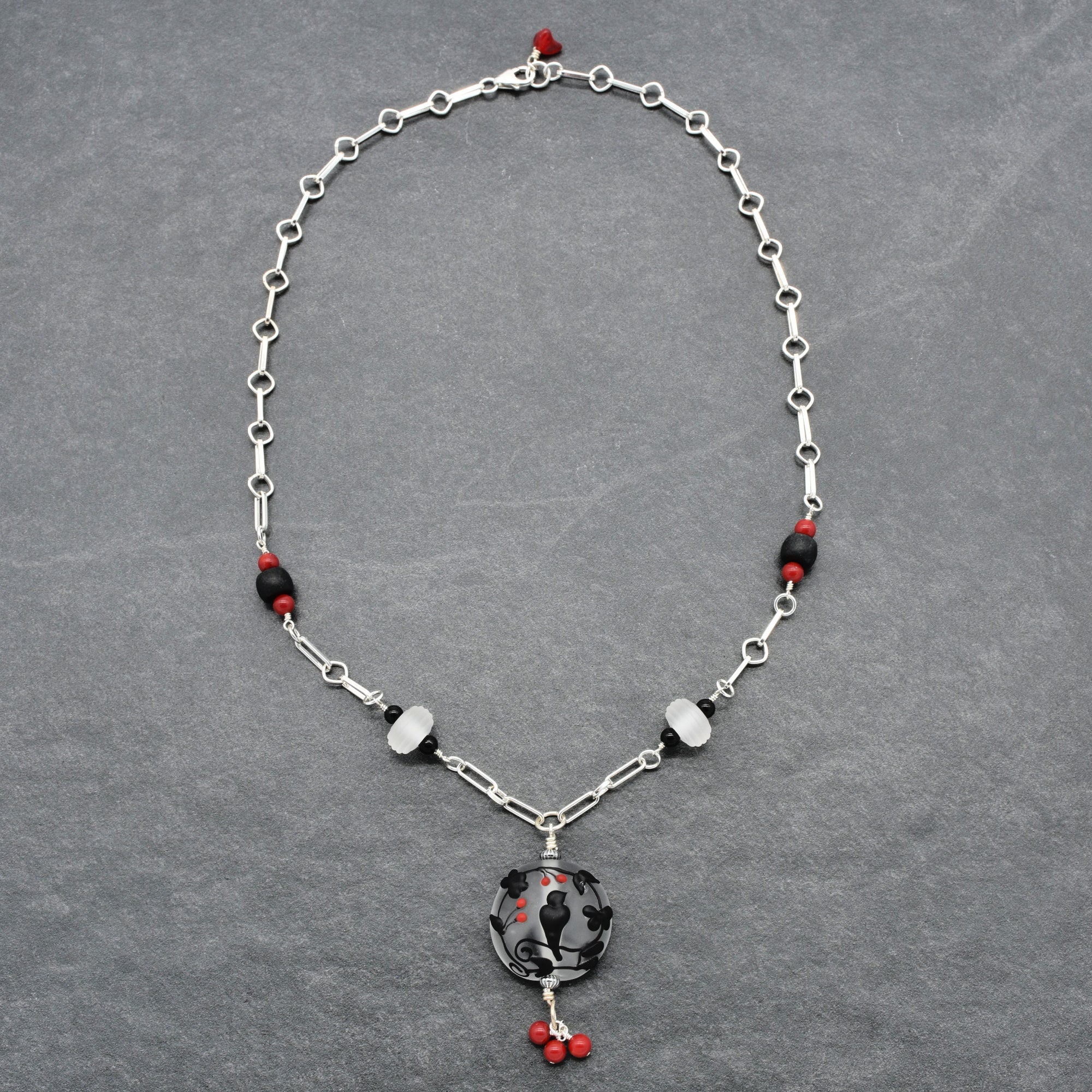A silver-toned beaded necklace with a circular black and red pendant, displayed on a textured gray background - made with love in Colorado by Ruby Acorn.