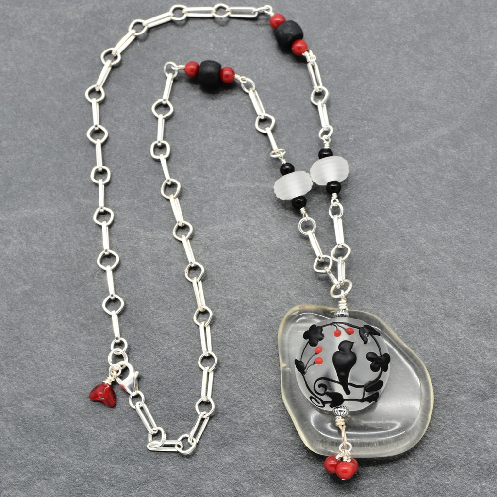 A silver link chain necklace featuring a lampwork glass pendant with a black and red floral design, accented with black and red beads on a gray textured background - made with love in Colorado by Ruby Acorn.