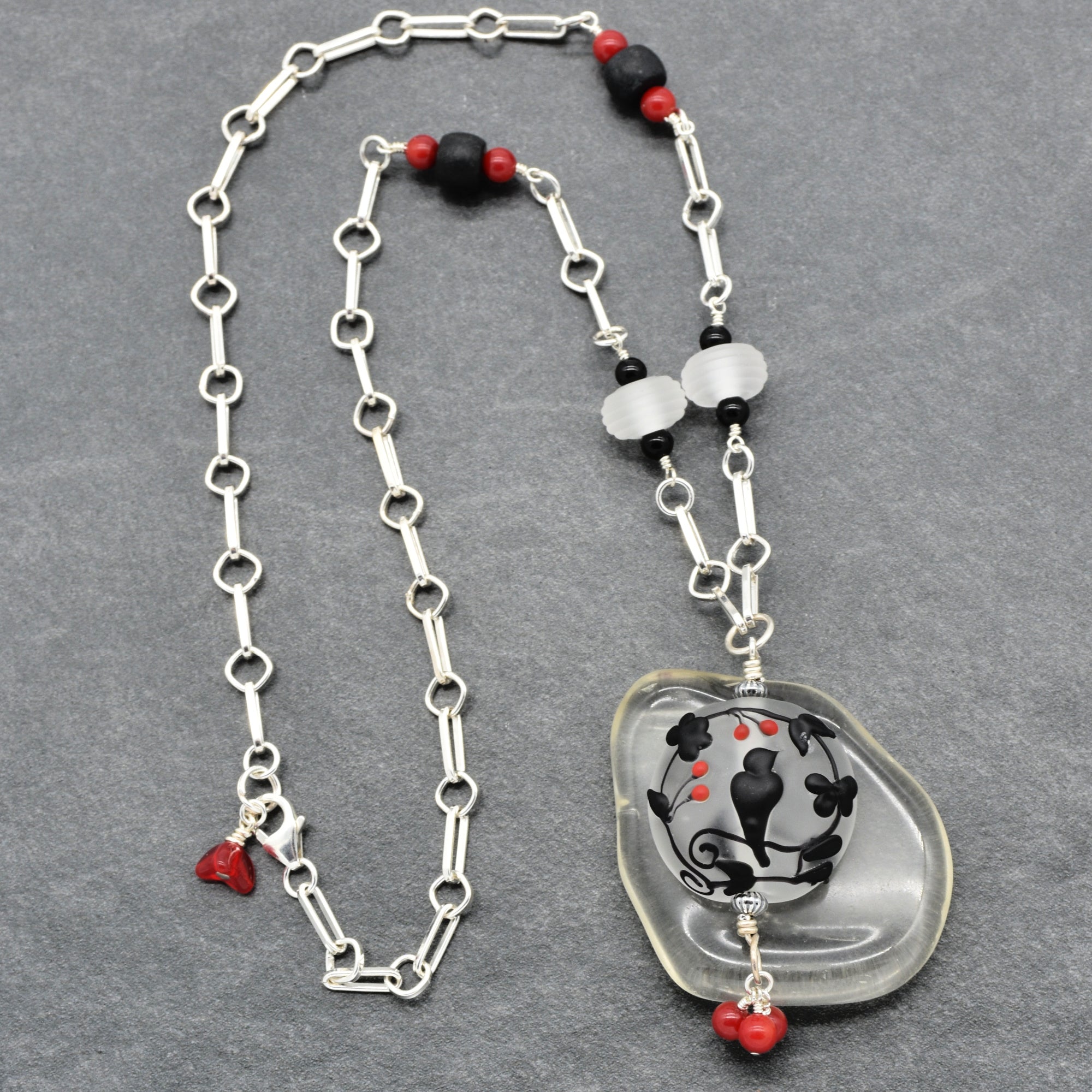 A silver link chain necklace featuring a lampwork glass pendant with a black and red floral design, accented with black and red beads on a gray textured background - made with love in Colorado by Ruby Acorn.