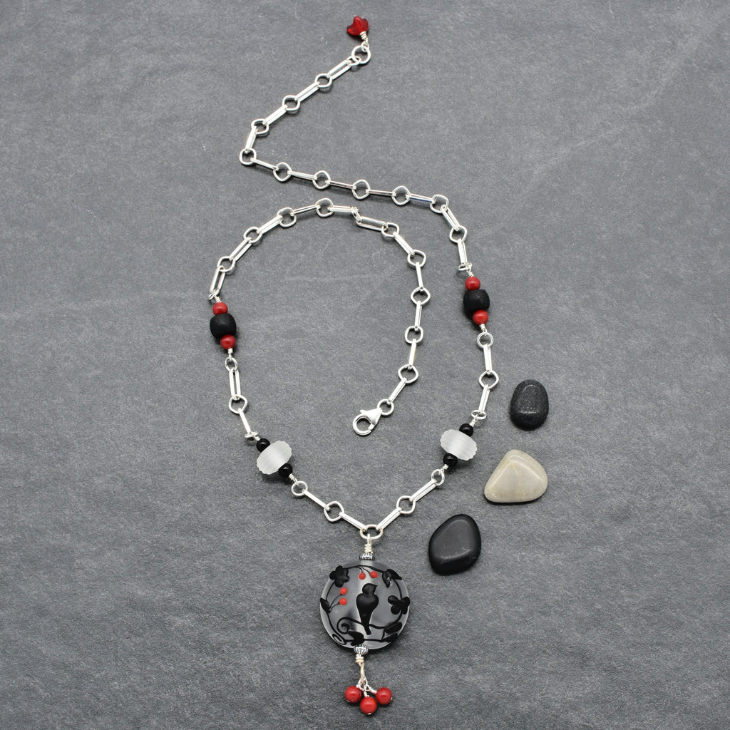 A silver-toned chain necklace with red and black beads and a black raven pendant with red accents, displayed on a textured gray background - made with love in Colorado by Ruby Acorn.