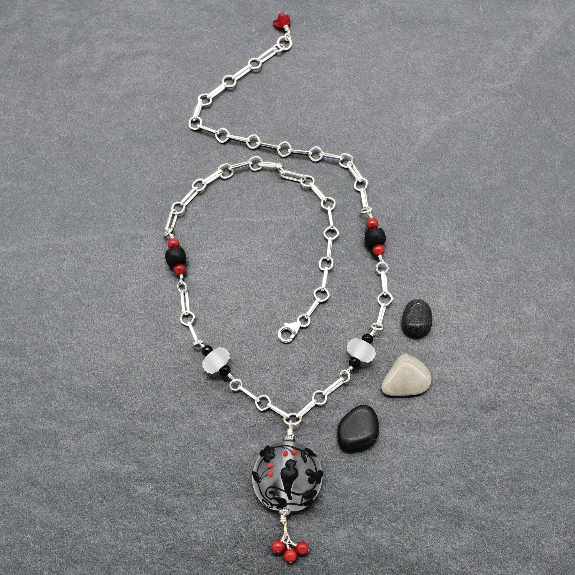 A silver-toned chain necklace with red and black beads and a black raven pendant with red accents, displayed on a textured gray background - made with love in Colorado by Ruby Acorn.