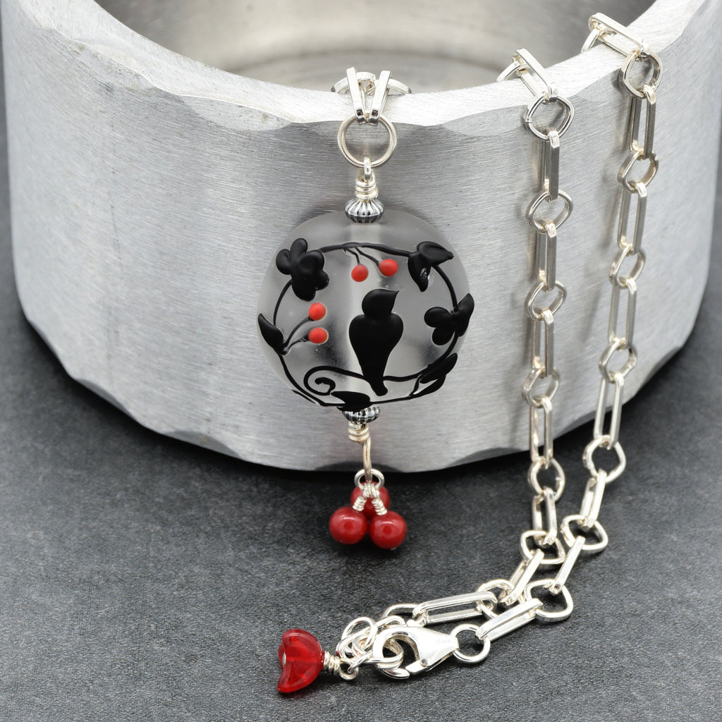 A silver chain necklace with a circular pendant featuring black bird and floral motifs over a gray background, accented with small red beads and a dangling red flower charm, displayed on a textured gray surface - made with love in Colorado by Ruby Acorn.