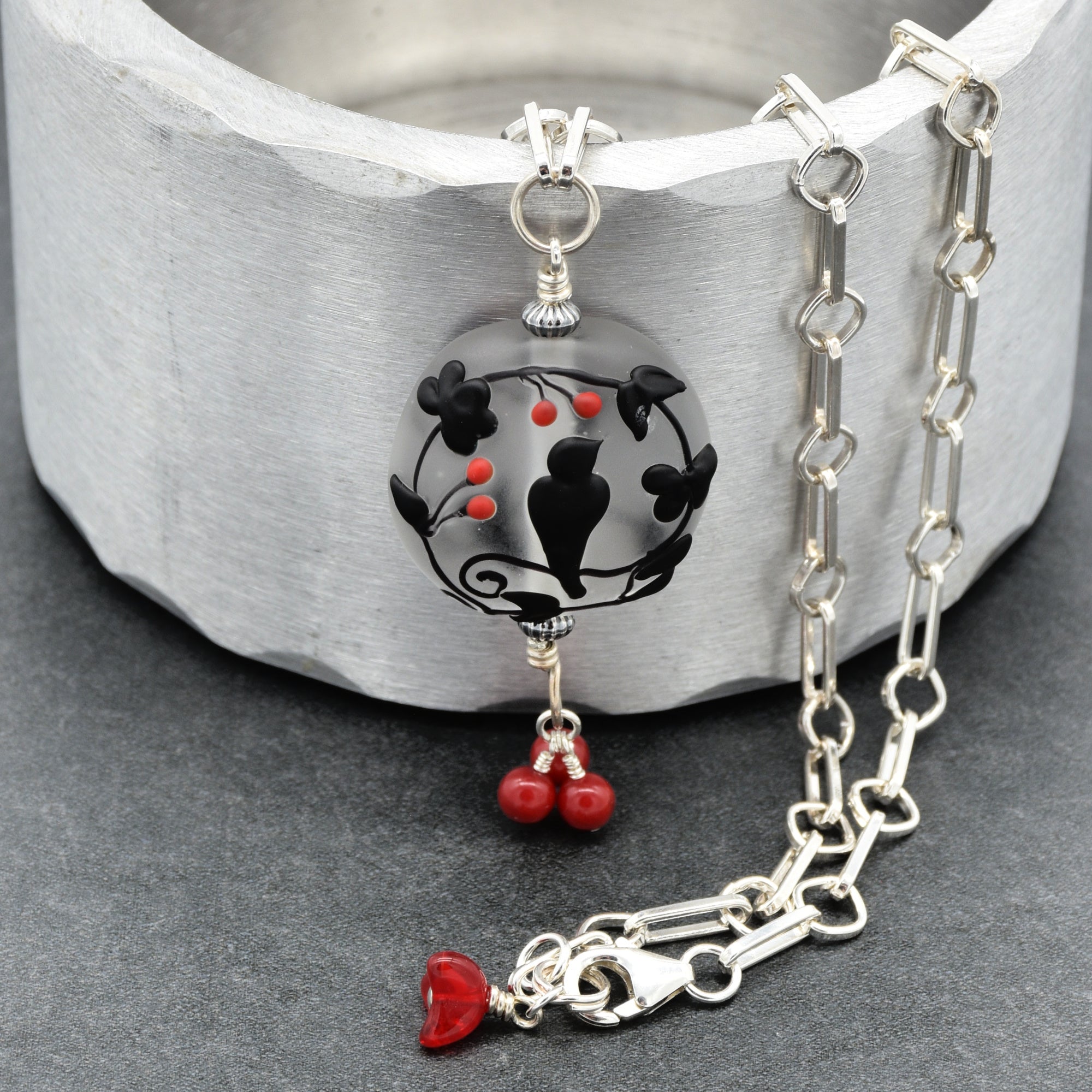A silver chain necklace with a circular pendant featuring black bird and floral motifs over a gray background, accented with small red beads and a dangling red flower charm, displayed on a textured gray surface - made with love in Colorado by Ruby Acorn.